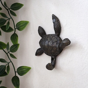 May include: A cast iron turtle wall hook with a brown finish. The hook is shaped like a turtle with its head and legs raised. The turtle's shell has a detailed texture.
