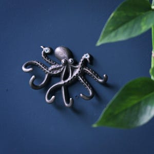 Könnte beinhalten: Ein bronzefarbener Oktopus-Wandhaken mit blauen Kristallaugen und blauen Akzenten an den Tentakeln. Der Oktopus ist an einer dunkelblauen Wand befestigt, mit grünen Blättern im Hintergrund.