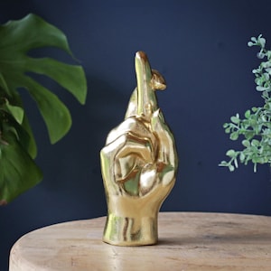 Könnte beinhalten: Eine goldfarbene Skulptur einer Hand mit gekreuztem Zeige- und Mittelfinger. Die Hand ist vertikal mit dem Handgelenk an der Basis positioniert und weist eine glatte, reflektierende metallische Oberfläche auf, die das Licht einfängt.