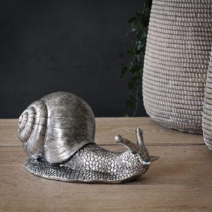 以下が含まれることがあります： 詳細な殻と質感のある体を持つ、銀色のカタツムリの置物。 カタツムリは木製の表面に置かれ、背景には織りのバスケットと緑があります。 この置物は装飾品です。