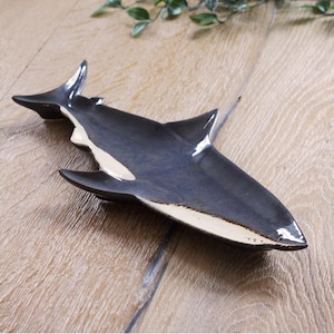 Può includere: Un piatto da portata in ceramica a forma di orca. Il piatto è principalmente blu scuro con dettagli bianchi e crema. Il piatto a forma di balena ha un design elegante e aerodinamico, perfetto per servire antipasti o piccoli piatti.