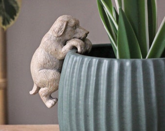 May include: A ceramic planter with a green glaze and a ribbed texture. A small, beige ceramic dog figurine is perched on the rim of the planter, with its paws gripping the edge.