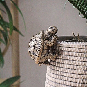 Puede incluir: Una figura de tortuga de color bronce agarrada al borde de una maceta tejida. La tortuga tiene un caparazón detallado con patrones geométricos y está colocada como si estuviera trepando. La maceta tiene una superficie texturizada y de color claro.