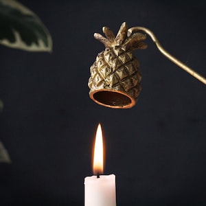 Op de afbeelding: Een gouden kaarsdover in de vorm van een ananas met een lange metalen handgreep. De dover is boven een witte kaars geplaatst.