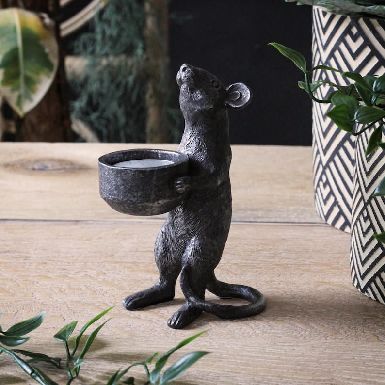 May include: A dark gray, textured mouse figurine standing on its hind legs. The mouse is holding a small, round, bowl-shaped candle holder in its front paws. The surface of the mouse and the bowl has a weathered, metallic appearance with visible details.