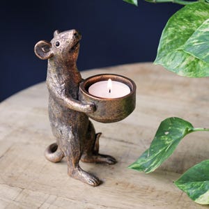 May include: A bronze-colored mouse figurine standing on its hind legs, holding a small, round bowl that contains a lit white tea light candle. The mouse has a textured, metallic finish and is looking upward with its ears perked and tail curled.