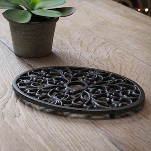 Op de afbeelding: Een ovale gietijzeren onderzetter met een ingewikkeld bloemmotief. De onderzetter heeft drie poten.