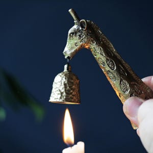 May include: A brass giraffe-shaped candle snuffer with a bell-shaped extinguisher. The giraffe's neck and head are detailed with textured patterns. A lit candle is positioned below the bell, with a dark blue background.