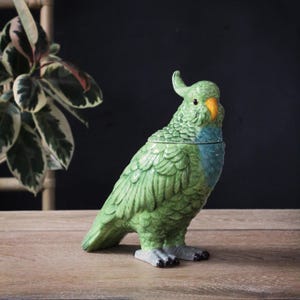 May include: A green ceramic parrot ornament with a blue chest and orange beak, standing on a wooden surface. The parrot has detailed feather patterns and a crest. It is a decorative item, possibly a container or a decorative piece for the home.
