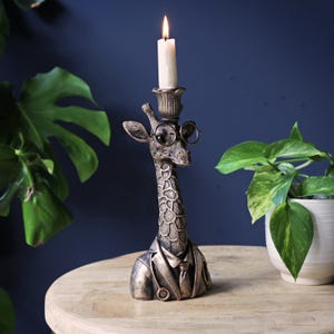 May include: A bronze-toned giraffe candle holder featuring a bust of a giraffe wearing a suit, tie, and round spectacles. The giraffe has a textured coat with dark brown spots and a candle cup on its head holding a lit white taper candle.