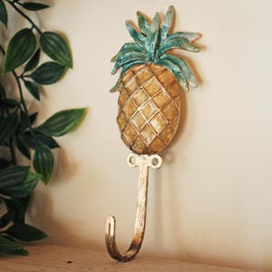 Peut inclure: Un crochet mural décoratif en forme d'ananas. L'ananas est doré avec un motif quadrillé, des feuilles vertes et un crochet doré. Le crochet est fixé sur un mur beige. La longueur du crochet est d'environ 15 cm.