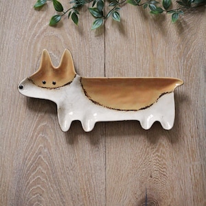 May include: A ceramic corgi-shaped dish with a brown and white color scheme. The dish has a long, rectangular body with four small legs and a raised head and ears. The eyes are small black dots. The dish is designed to hold small items.