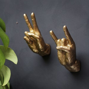 Op de afbeelding: Twee gouden metalen handbeelden, een maakt een vredesteken en de andere maakt een rock-and-roll handgebaar.