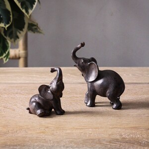 May include: Two small, dark brown elephant figurines. One is sitting, the other is standing with its trunk raised. The figurines are on a light brown wooden surface, with a plant in the background.