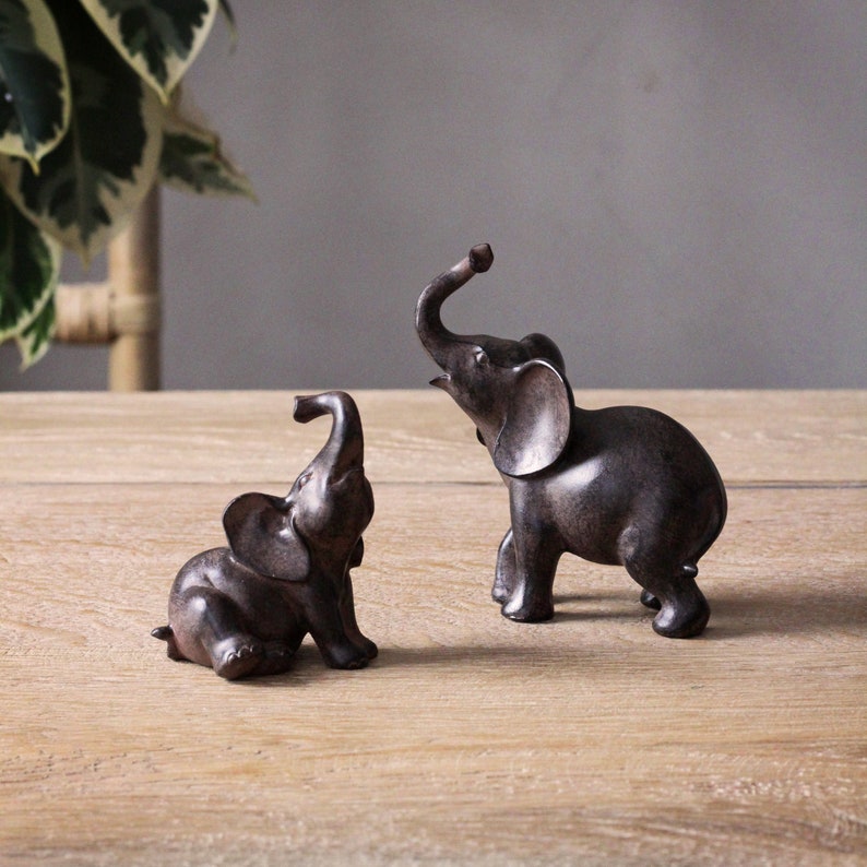 May include: Two small, dark brown elephant figurines. One is sitting, the other is standing with its trunk raised. The figurines are on a light brown wooden surface, with a plant in the background.