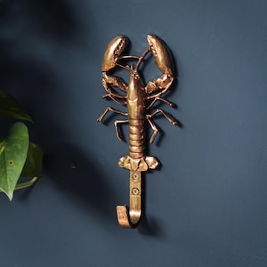 May include: A metallic copper-coloured wall hook shaped like a lobster. The lobster figure features detailed claws, legs, and a segmented body. A functional J-shaped hook extends from the tail, secured to the wall with two visible screws.