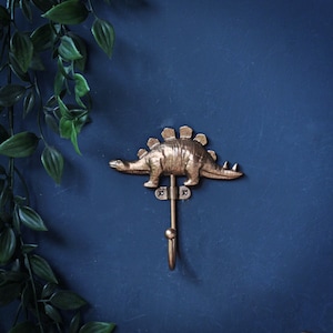 May include: A metallic gold-toned wall hook shaped like a Stegosaurus dinosaur. The dinosaur figure is mounted on a small rectangular plate with two visible screws, and a curved hook extends downward from the center of the dinosaur's body.