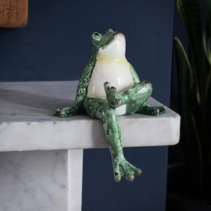 May include: A green ceramic frog figurine with a white belly, sitting on a white marble surface with one leg dangling off the edge. The frog has a yellow stripe around its neck and is looking up.