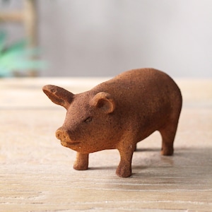 Op de afbeelding: Een klein, bruin keramiek varkensbeeldje. Het varken heeft een getextureerd oppervlak en staat op een houten oppervlak.