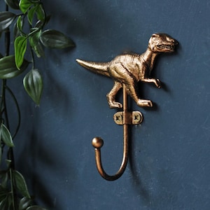 May include: A metallic bronze-colored wall hook featuring a sculpted dinosaur figure mounted above a curved hook. The piece is secured to the wall with a small metal plate and two visible screws, providing a functional storage solution.