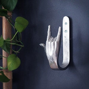 May include: Silver metal wall hook shaped like a hand giving the 'I love you' sign. The hook is mounted on a dark grey wall.