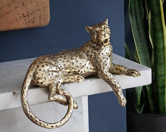 May include: A gold-toned leopard figurine with a textured, spotted surface. The leopard is depicted in a reclining position with its front paws extended over the edge and its long tail curled downward, showcasing a detailed feline facial expression.