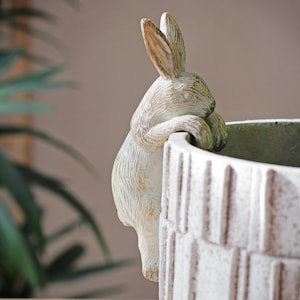 Pode incluir: Vaso de cerâmica branco com uma figura de coelho espiando por cima da borda. O vaso tem um design texturizado e nervurado.