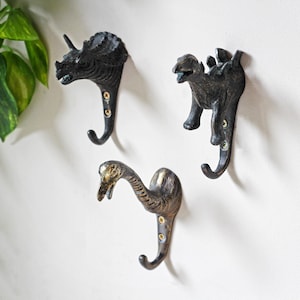 May include: Three wall-mounted hooks made of dark, metallic material. Each hook features a different dinosaur figure: a triceratops, a stegosaurus, and a long-necked sauropod. Each piece is secured to the wall with two visible gold-colored screws.