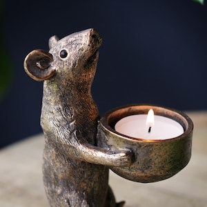 May include: A bronze-colored mouse figurine standing upright and holding a small, round, bowl-shaped candle holder. The mouse has textured fur details and a dark eye. A white tea light candle with a lit flame is placed inside the bowl holder.