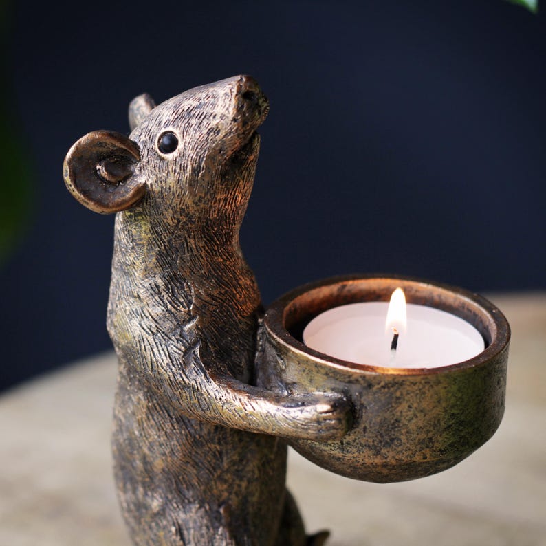 May include: A bronze-colored mouse figurine standing upright and holding a small, round, bowl-shaped candle holder. The mouse has textured fur details and a dark eye. A white tea light candle with a lit flame is placed inside the bowl holder.