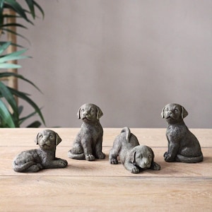 May include: Four small, gray, stone-like sculptures of dogs in various poses. The sculptures are arranged on a wooden surface.