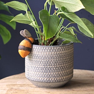May include: A cylindrical gray planter with a textured diamond pattern on the exterior. A small, orange and black bumblebee figurine is attached to the rim. A green leafy plant is potted inside the container, with its stems extending upward.