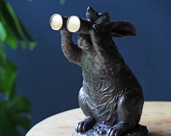 May include: A dark bronze rabbit ornament holding binoculars. The rabbit is in a seated pose, with its ears pricked. The binoculars have gold lenses. The ornament is on a wooden surface.