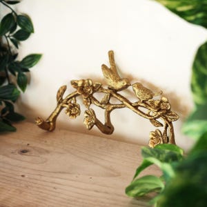 May include: A gold-coloured metal wall hook featuring a branch design with two birds perched on it. The branch has several hooks and is adorned with flowers. The hook is designed to be mounted on a wall.