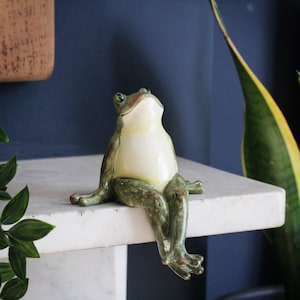 Puede incluir: Una figurita de cerámica de rana con pintura verde y blanca, sentada en un estante blanco. La rana tiene una amplia sonrisa y mira hacia arriba.
