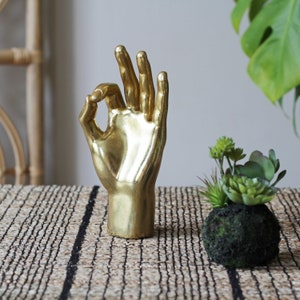 Può includere: Una scultura di mano dorata che fa il segno OK. La mano è su un runner da tavolo intrecciato con strisce nere.