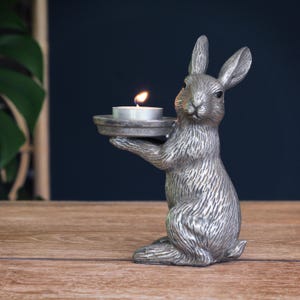 May include: A silver-colored rabbit figurine holding a small, round candle holder with a lit candle. The rabbit is in a seated position with its ears upright. The candle flame is orange. The figurine is on a wooden surface.