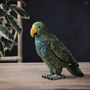 May include: A ceramic parrot figurine in shades of green and teal, with a yellow beak and black eyes. The bird has a textured, scale-like pattern and stands on a wooden surface. The figurine is a decorative item.