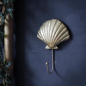 Puede incluir: Un colgador de pared con forma de concha dorada. El gancho de metal tiene un diseño curvo. La concha tiene una textura con un patrón acanalado. El gancho está montado en una pared azul oscuro, con vegetación en el fondo.