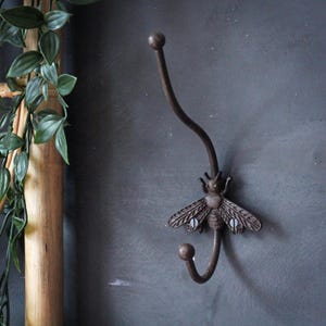 May include: A decorative wall hook in a dark bronze finish, featuring a stylized bee design. The hook curves downward, with a small ball at the end. The bee's wings are detailed, adding a touch of nature-inspired decor.