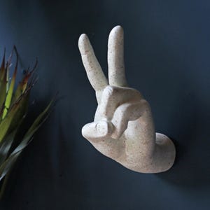 Könnte beinhalten: Eine beigefarbene, an der Wand befestigte Handskulptur, die das Friedenszeichen zeigt. Die Skulptur hat eine strukturierte Oberfläche mit kleinen braunen Sprenkeln. Der Hintergrund ist eine dunkelblaue Wand, mit einem Blick auf eine grüne Pflanze links.