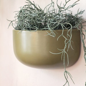 Może przedstawiać: Oliwkowo-zielona donica ścienna wypełniona zwisającymi, jasnozielonymi roślinami. Donica ma zaokrąglone dno i kwadratową górę, tworząc nowoczesną, minimalistyczną estetykę. Delikatne łodygi rośliny opadają w dół, dodając naturalnego charakteru.