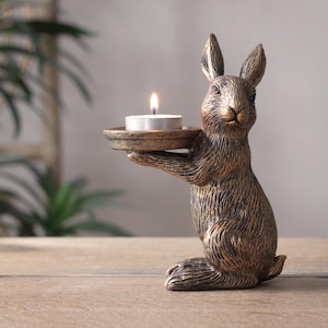 May include: A gold-toned rabbit figurine holding a small, round candle holder. The rabbit is standing on a wooden surface and has a textured fur design. The candle holder is empty.