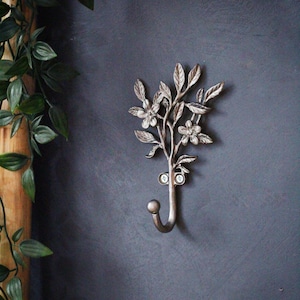 Puede incluir: Un gancho de pared decorativo de metal plateado con forma de rama floreciente. El gancho tiene hojas y flores detalladas. Está montado en una pared gris oscuro, con vegetación a la izquierda.