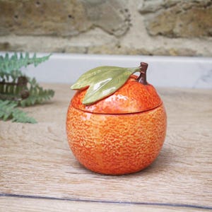Könnte beinhalten: Ein keramikfarbener, orangenförmiger Behälter mit Deckel und grünen Blättern. Der Behälter hat eine leuchtend orange Farbe mit einer strukturierten Oberfläche. Die Blätter sind glänzend grün. Der Behälter steht auf einer Holzoberfläche.