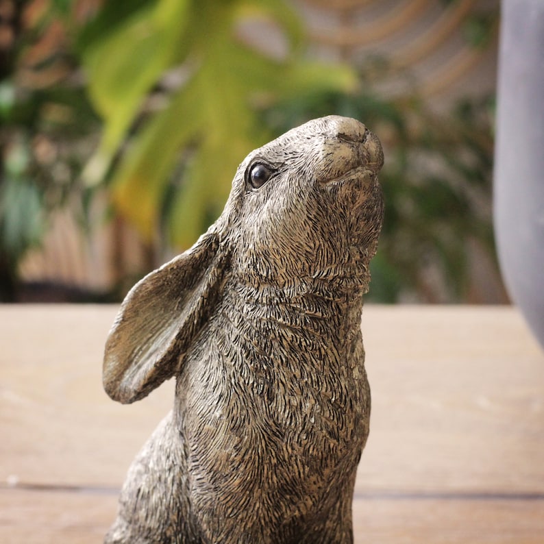 May include: A bronze-effect rabbit sculpture, with detailed fur texture and focused eyes looking upwards. The statue is set against a blurred backdrop of green foliage and a neutral surface, suitable for a garden or interior setting.