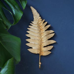 May include: A gold-colored metal wall hook shaped like a fern leaf. The hook features a detailed, textured leaf design with a central stem that extends downward into a curved hook at the base, providing a functional element for hanging items.