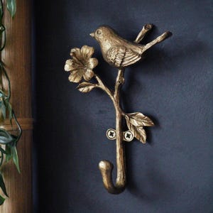 Puede incluir: Un gancho de pared decorativo de metal dorado con forma de pájaro posado en una rama con una flor y hojas. El gancho está montado en una pared azul oscuro. El pájaro y la rama están detallados.