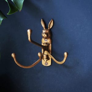 Puede incluir: Un gancho de pared de metal color latón con una cabeza de conejo esculpida en la parte superior. El accesorio incluye tres brazos curvos y articulados que se extienden hacia afuera, cada uno terminando en una punta redondeada.