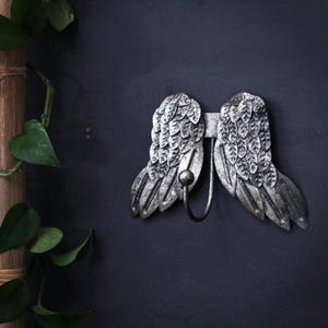 Puede incluir: Un gancho de pared de metal con un par de alas de ángel plateadas y texturizadas. Las alas presentan patrones de plumas superpuestas y un gancho central curvado con una punta esférica, ofreciendo una solución de almacenamiento funcional.